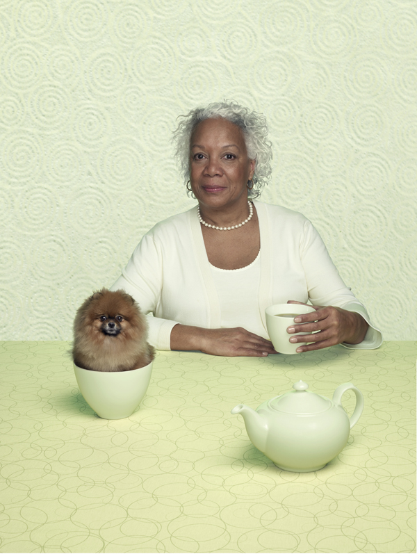A teacup pomeranian sits in a teacup while an elegant older woman drinks green tea. 